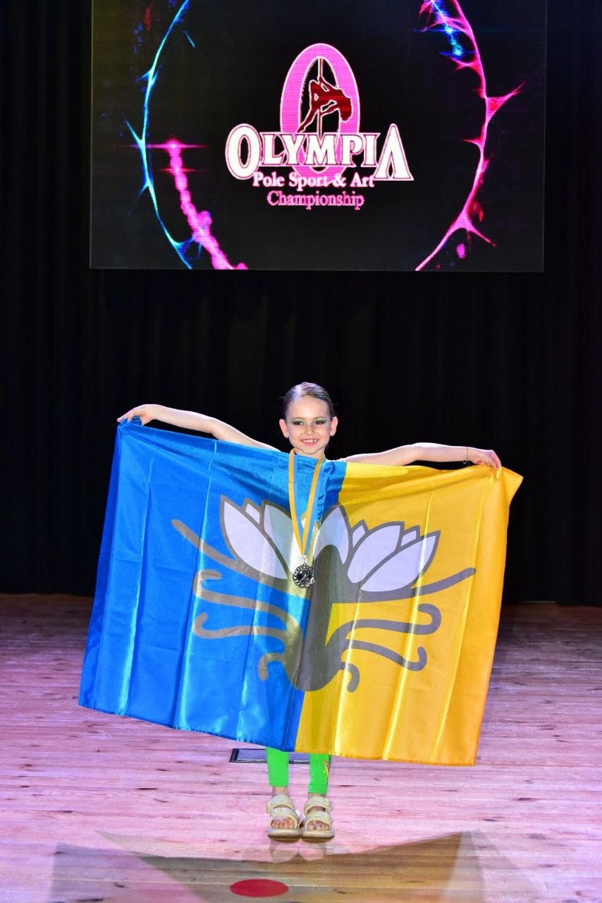 Юна спортсменка з медаллю на шиї стоїть на сцені та тримає за спиною великий синьо-жовтий прапор із малюнком латаття. На задньому плані видно темну завісу та екран із логотипом змагань «Olympia Pole Sport & Art Championship».