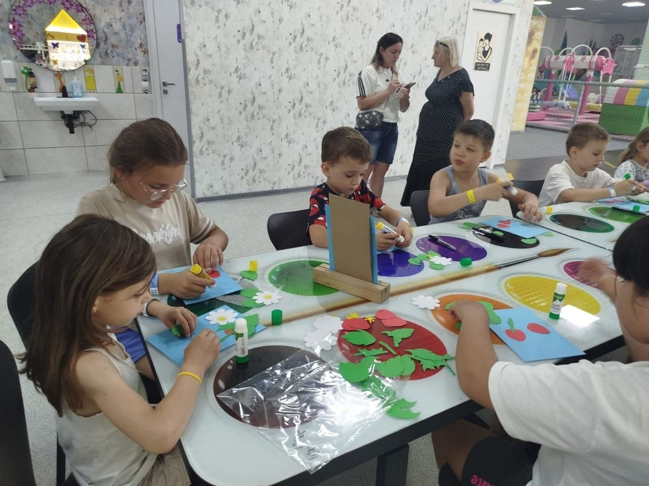 Група дітей сидить за великим столом у світлій кімнаті та займається творчістю, створюючи аплікації з кольорового паперу та клею. На задньому плані стоять дві дорослі жінки, а поруч видно ігрову зону.