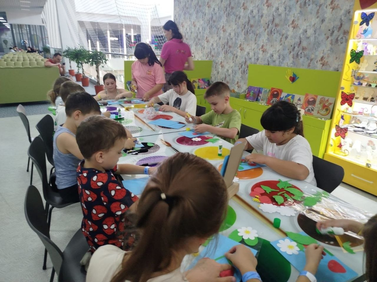 Діти сидять уздовж довгого столу у світлій дитячій кімнаті, захоплено майструючи паперові аплікації та вироби з клеєм. На задньому плані видно дорослу жінку у рожевій футболці, зелені меблі та яскраву вітрину з декором.