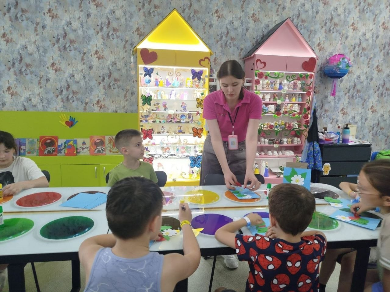 Аніматор у рожевій футболці допомагає дітям за великим столом створювати паперові аплікації. На задньому плані кімнати стоять дві яскраві шафи-вітрини у формі будиночків із дрібним декором на полицях.