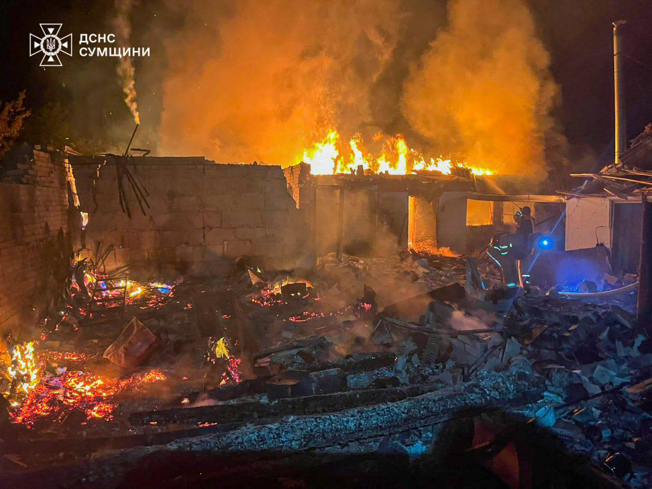 Рятувальники ліквідовують масштабну нічну пожежу, яка охопила зруйновану будівлю, де палають стіни та дах серед уламків. У лівому верхньому кутку розміщено емблему та напис «ДСНС СУМЩИНИ».