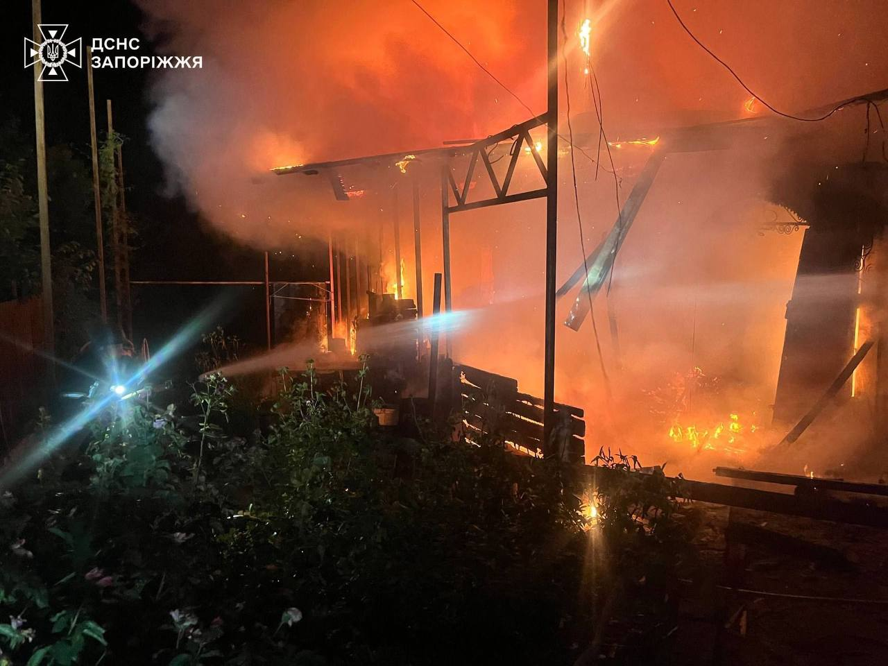 Рятувальники гасять потужну нічну пожежу, що охопила приватну будівлю, яка палає яскравим вогнем серед густого диму. У лівому верхньому кутку розміщено емблему та напис «ДСНС ЗАПОРОЖЖЯ».
