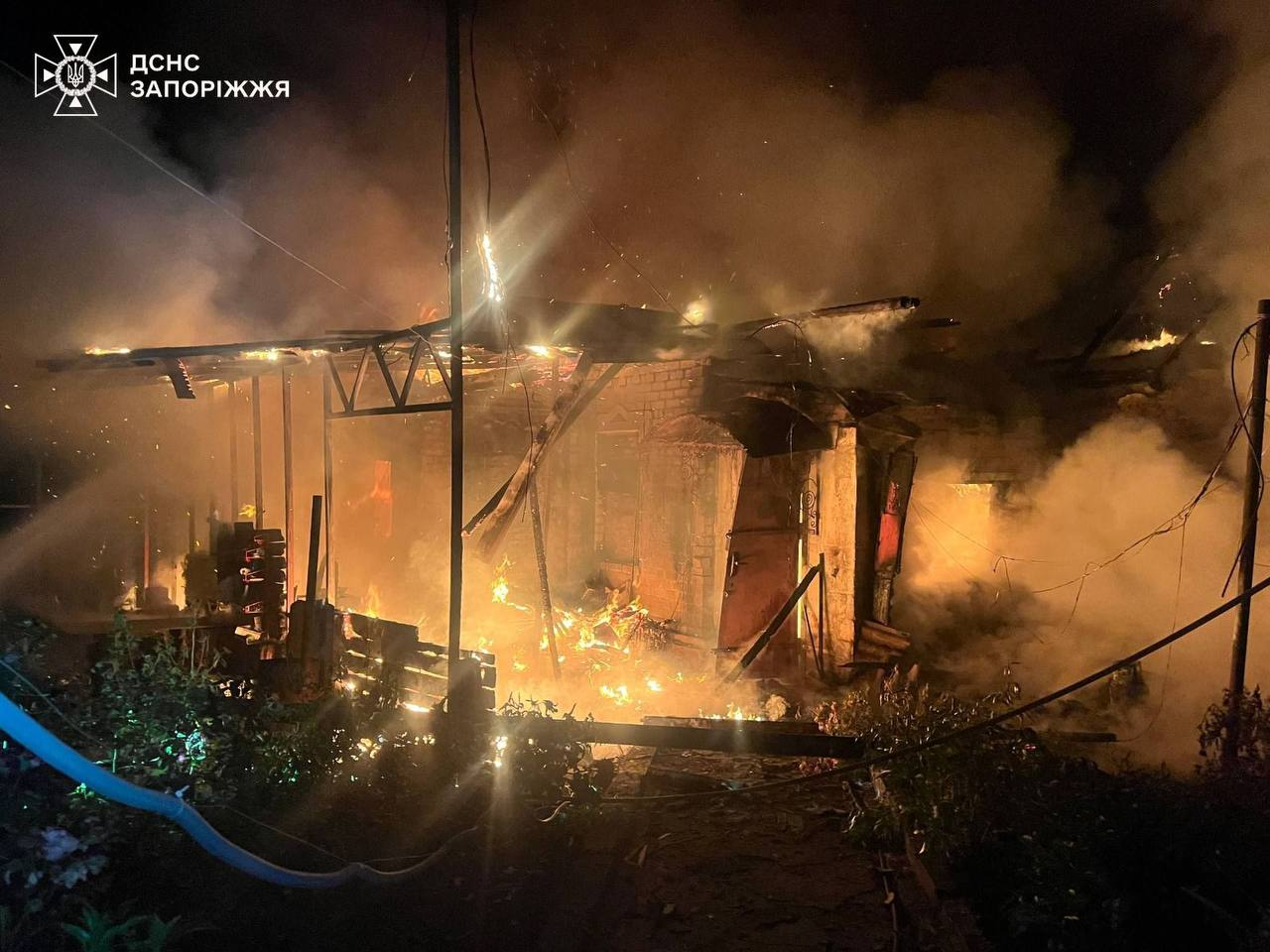Ліквідація нічної пожежі в приватному секторі, де відкрите полум'я та густий дим охопили зруйновану металеву та цегляну конструкцію будівлі. У лівому верхньому кутку розміщено логотип із написом «ДСНС ЗАПОРОЖЖЯ».