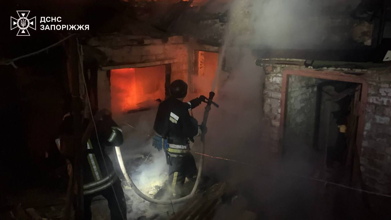Рятувальники в захисному екіпіруванні гасять пожежу всередині зруйнованої будівлі, спрямовуючи струмінь води у задимлене приміщення. У лівому верхньому кутку розміщено емблему та напис «ДСНС ЗАПОРОЖЖЯ».