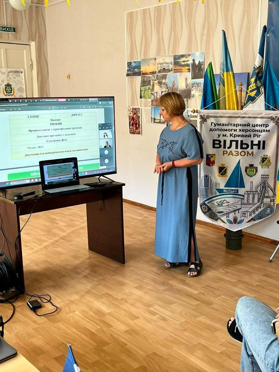 Жінка у довгій блакитній сукні проводить презентацію в приміщенні гуманітарного центру, стоячи біля великого екрана з переліком документів. Поруч розміщено прапори України та банер «Гуманітарний центр допомоги херсонцям у м. Кривий Ріг «Вільні разом».