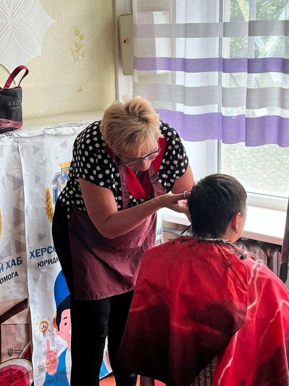 Жінка-перукар у блузі в горошок та фартуху стриже клієнта, накритого червоним пеньюаром, біля вікна в приміщенні. На задньому плані видно частину банера з написом «ХЕРСОН» та сумку на полиці.