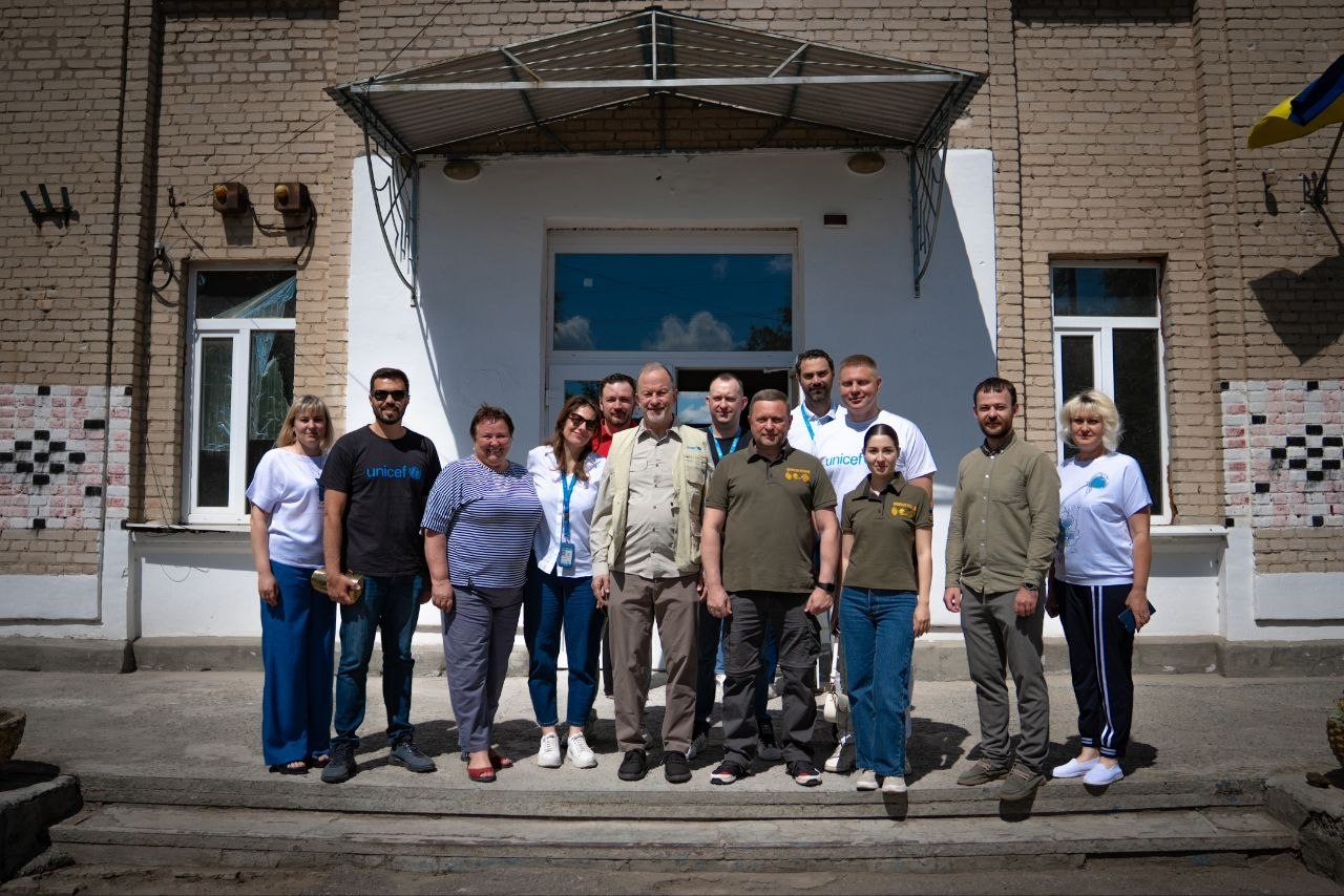 Група людей стоїть у ряд для спільного фото біля центрального входу до цегляної будівлі під навісом. Кілька осіб одягнені у білі та чорні футболки з логотипом UNICEF, а у верхньому правому кутку будівлі видно частину прапора України.