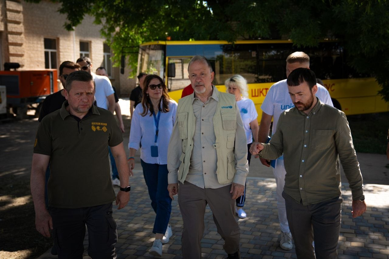 Група людей у цивільному одязі та брендованих речах UNICEF іде брукованою доріжкою на подвір'ї будівлі. На задньому плані видно зелені дерева та припаркований жовтий автобус.