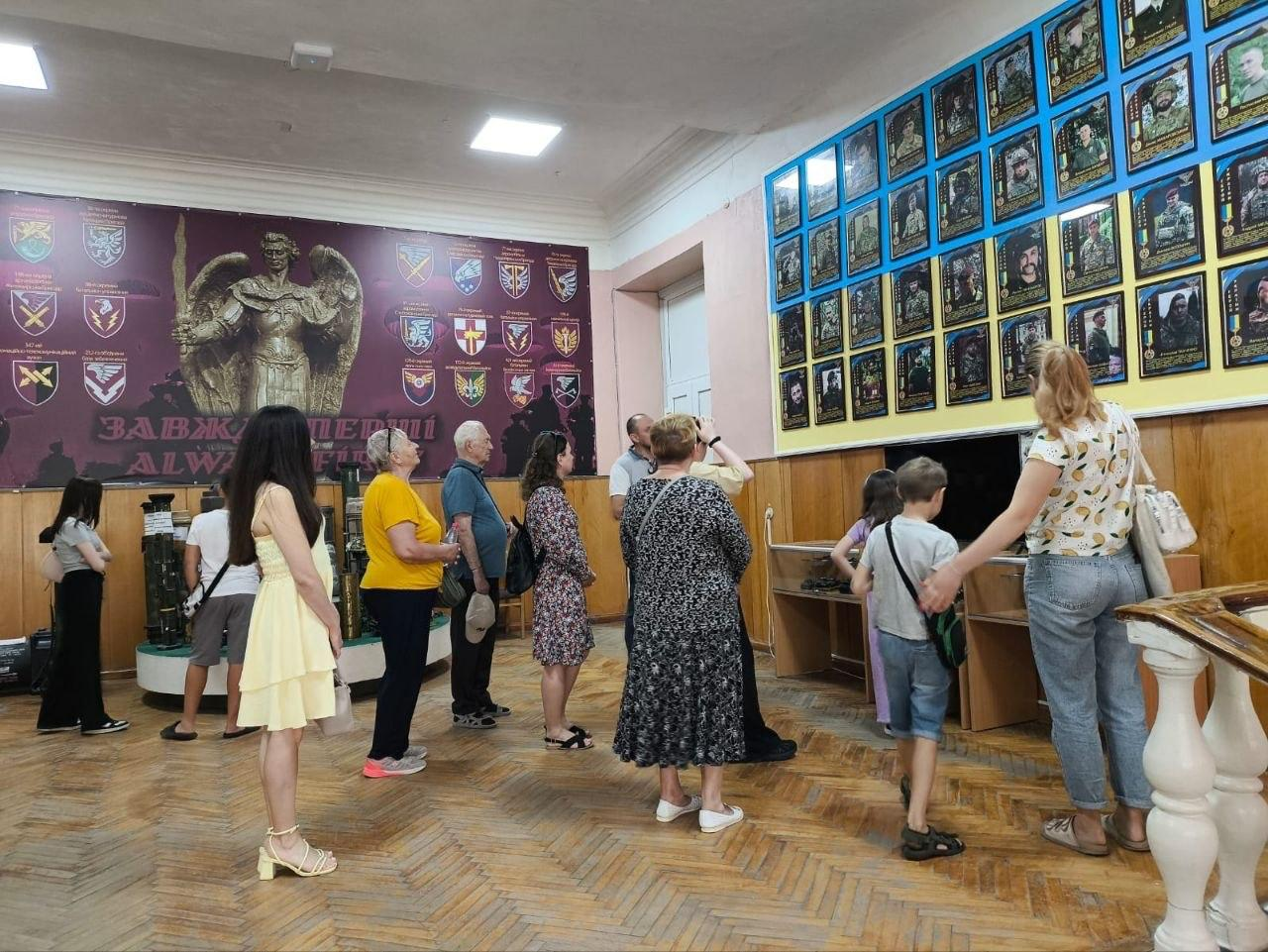 Група відвідувачів різного віку розглядає експозицію у музейній залі з паркетною підлогою. На стінах розміщені великий темний банер із військовою символікою та пам'ятна стіна з численними портретами у рамках.