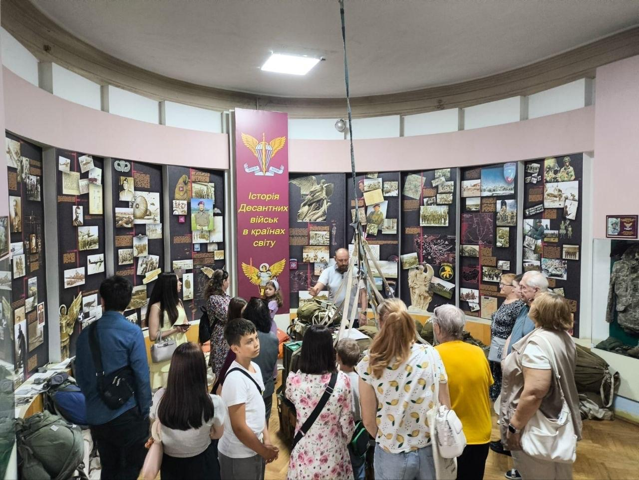 Екскурсовод проводить презентацію для групи відвідувачів у напівкруглій музейній залі, присвяченій історії десантних військ. Люди стоять навколо центральної експозиції з парашутним спорядженням на тлі стін, що повністю завішені інформаційними стендами, фотографіями та плакатами.