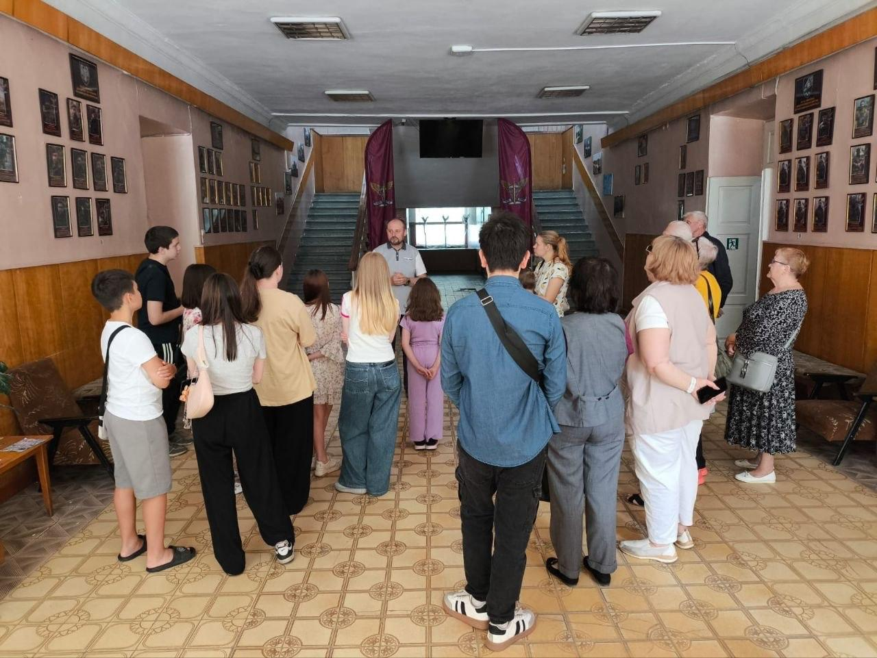 Група відвідувачів стоїть півколом у просторому холі будівлі та слухає екскурсовода, який стоїть попереду біля сходів на другий поверх. На стінах коридору розміщені численні ряди невеликих портретів у рамках, а підлога викладена світлою плиткою з візерунком.