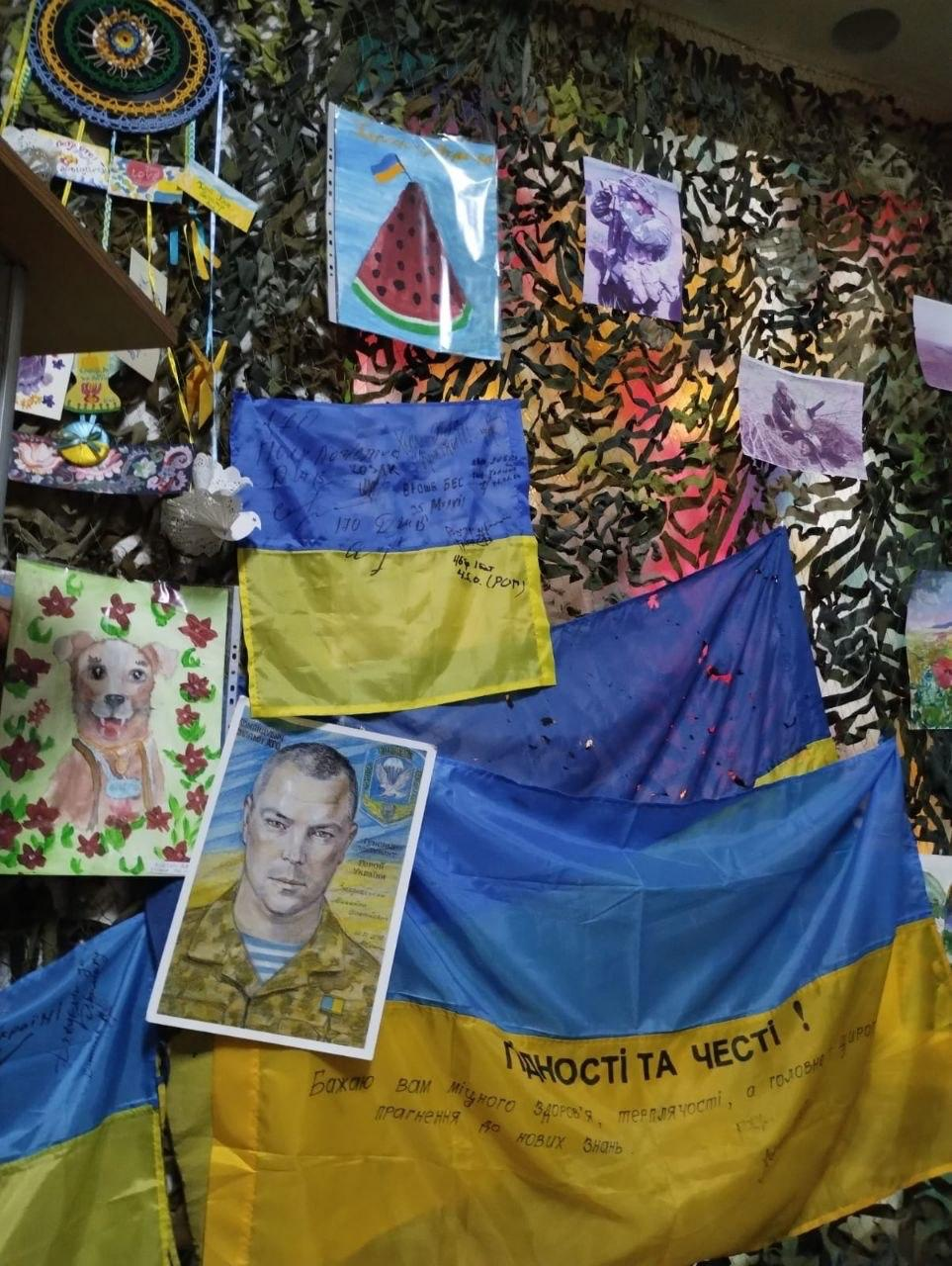 Патріотичний куточок пам'яті, оформлений на тлі маскувальної сітки. На стіні закріплені синьо-жовті прапори з написами-побажаннями, портрет військовослужбовця в однострої, дитячі малюнки собаки та кавуна, а також напис «Гідності та честі!».