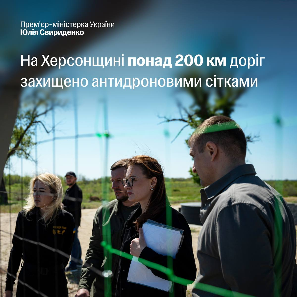 Група людей у цивільному одязі та військовій формі стоїть на вулиці та спілкується, дивлячись крізь металеву антидронову сітку. У верхній частині зображення міститься білий текст про захист понад 200 км доріг на Херсонщині антидроновими сітками.