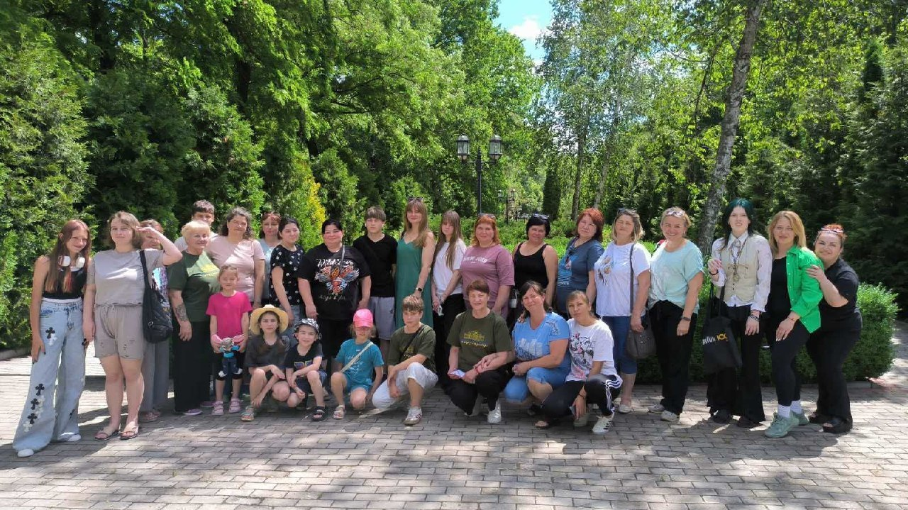 Велика група дітей та дорослих позує для спільного фото на брукованій доріжці в парку. Вони стоять і сидять на колінах на тлі густих зелених дерев, кущів та ліхтарного стовпа.