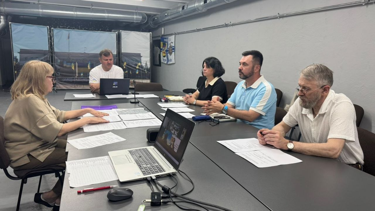 П'ятеро людей сидять навколо Т-подібного столу у сірому робочому кабінеті під час засідання.