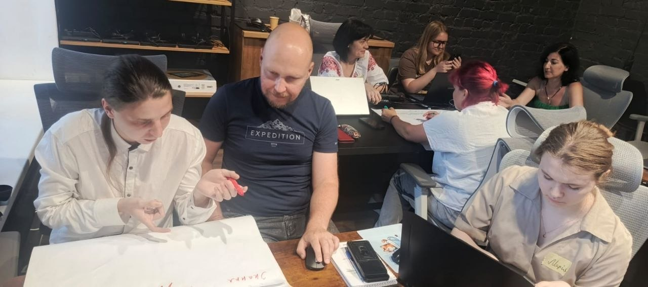 Група людей сидить за великим столом в офісі або навчальній аудиторії під час спільної роботи, обговорення чи тренінгу, використовуючи ноутбуки та паперові матеріали.