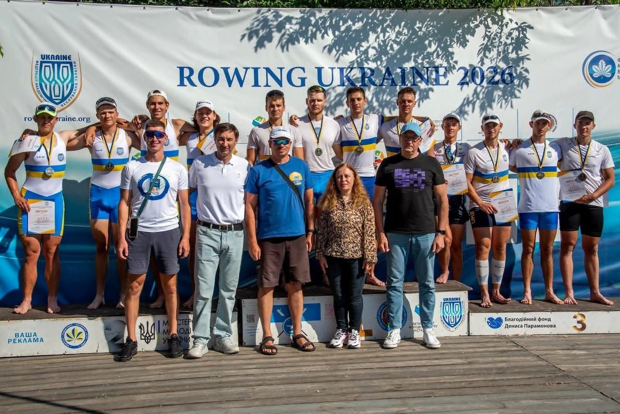 Група спортсменів-веслувальників та їхніх тренерів стоїть на п'єдесталі під час церемонії нагородження на тлі офіційного банера «ROWING UKRAINE 2026».