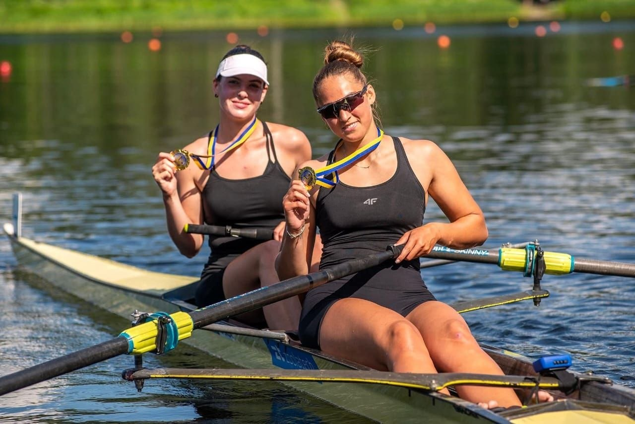 Дві спортсменки в чорній формі сидять у човні для академічного веслування на воді та тримають у руках завойовані золоті медалі.