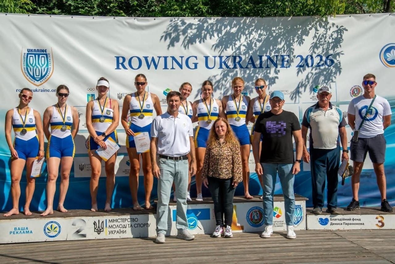 Група з восьми спортсменок-веслувальників у синьо-жовтій формі та їхніх тренерів стоїть на п'єдесталі під час церемонії нагородження на тлі офіційного банера «ROWING UKRAINE 2026».