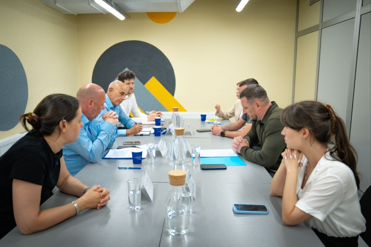 Група людей сидить за довгим столом під час ділової зустрічі або переговорів у світлому офісному кабінеті.