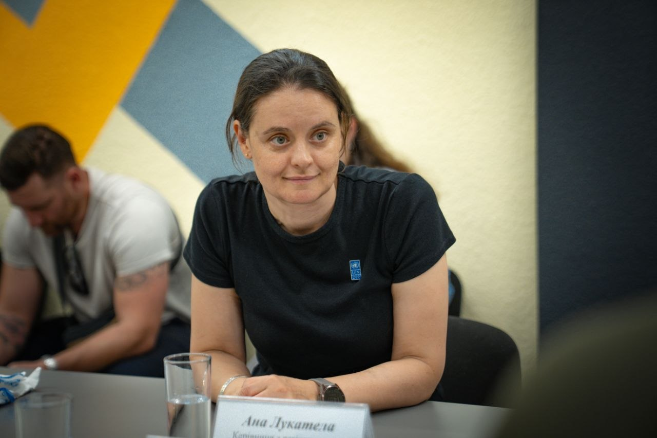 Жінка у чорній футболці з логотипом ПРООН (UNDP) сидить за столом під час ділової зустрічі.