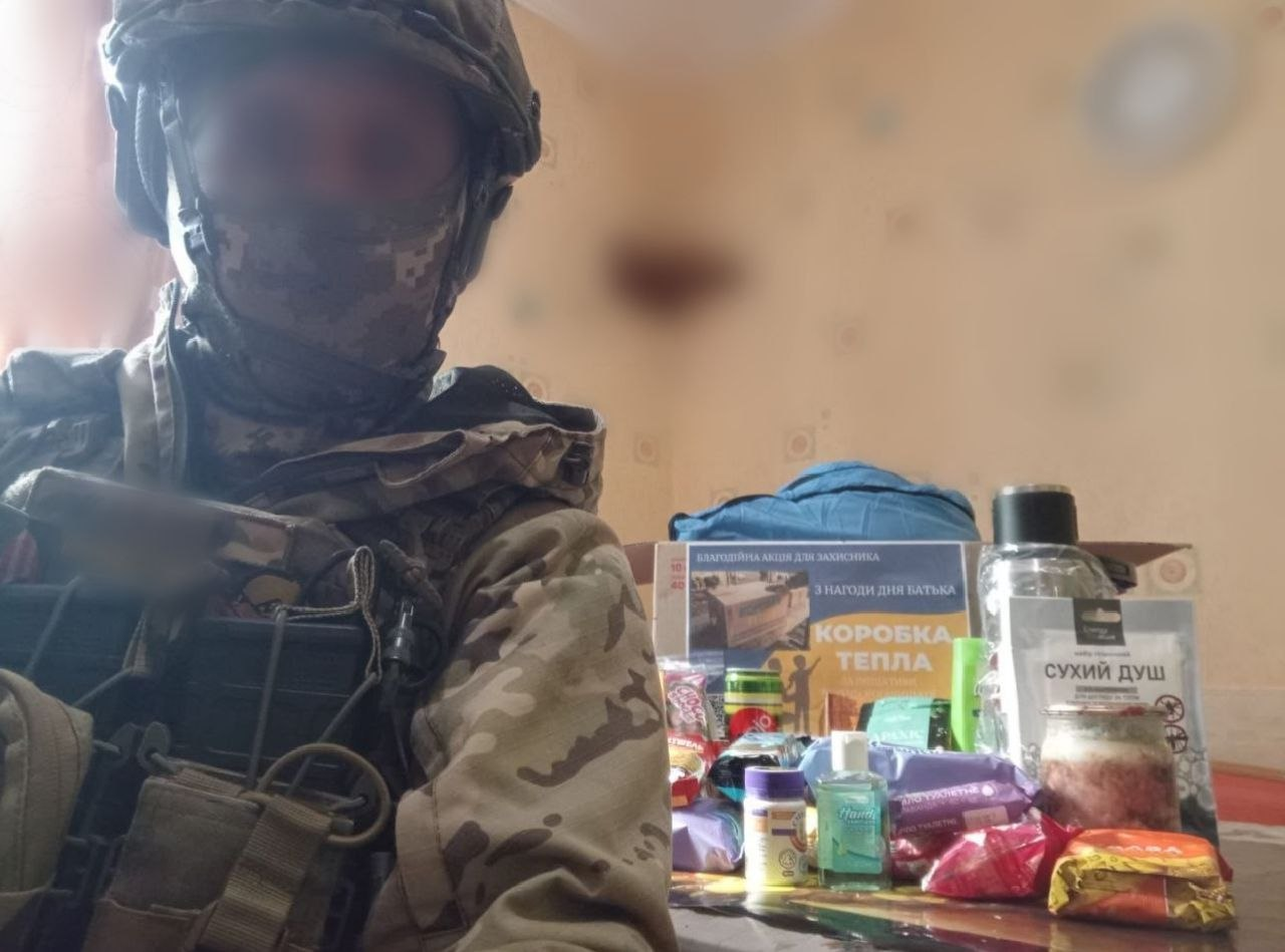 Військовослужбовець у повному екіпіруванні та касці стоїть поруч із благодійною допомогою, серед якої коробка з написом «Коробка тепла», сухий душ, засоби гігієни та продукти харчування.