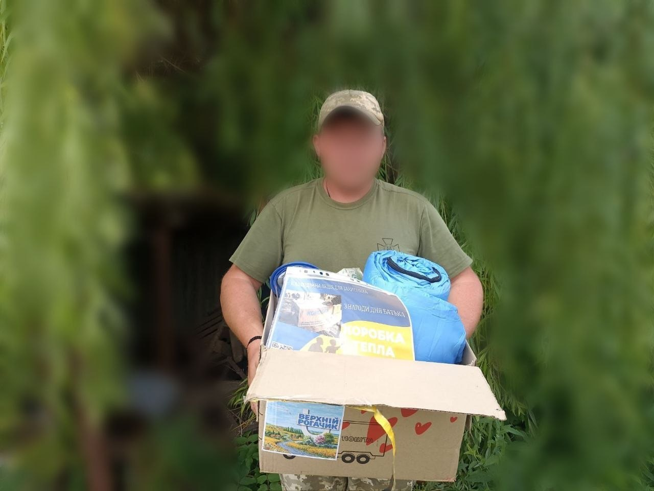 Чоловік у камуфляжній кепці та футболці хакі тримає велику картонну коробку з гуманітарною допомогою, де видно спальні мішки та пакунок із написом «Коробка тепла», на фоні зелених кущів.