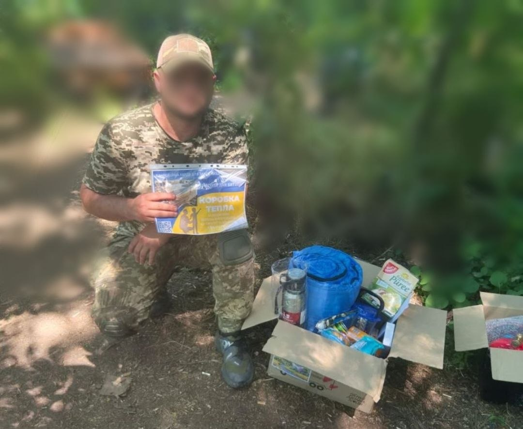 Військовослужбовець у камуфляжній формі сидить навпочіпки на землі та тримає листівку з написом «Коробка тепла» поруч із відкритими коробками з гуманітарною допомогою серед дерев.