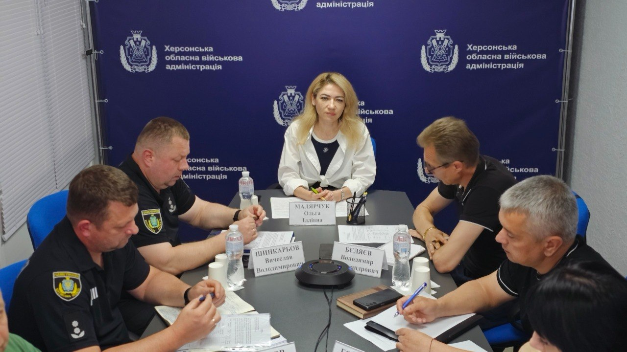 Робоча зустріч посадовців та представників служб за столом у кабінеті на тлі синього брендованого банера з гербом.