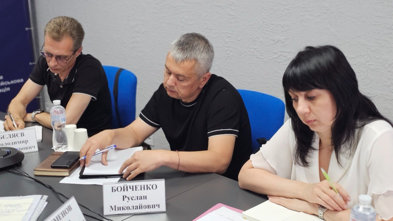 Троє учасників робочої наради — двоє чоловіків та жінка — сидять за столом з документами та роблять нотатки.