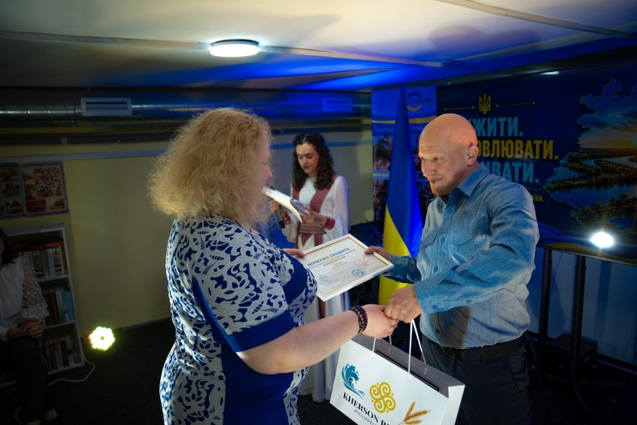 Чоловік у синій сорочці та темних штанах вручає почесну грамоту та подарунковий пакет жінці у синьо-білій сукні під час урочистого заходу на тлі Державного Прапора України.