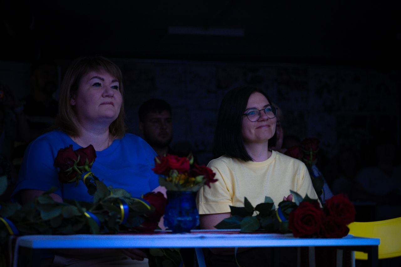 Дві жінки — одна в синій кофті, інша в жовтій футболці та окулярах — сидять за столом у напівтемній залі поруч із букетами червоних троянд під час урочистого заходу.