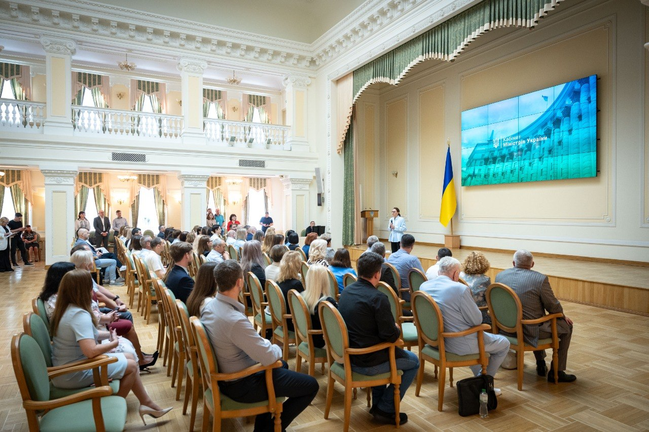 Великий світлий зал із колонами та балконами, де на стільцях сидить багато людей, які слухають доповідача біля великого екрана з написом «Кабінет Міністрів України».