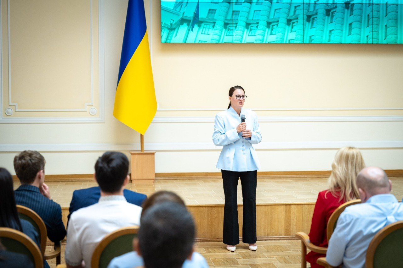 Жінка в окулярах, світло-блакитному жакеті та чорних штанах виступає з мікрофоном на сцені перед аудиторією на тлі державного прапора України.