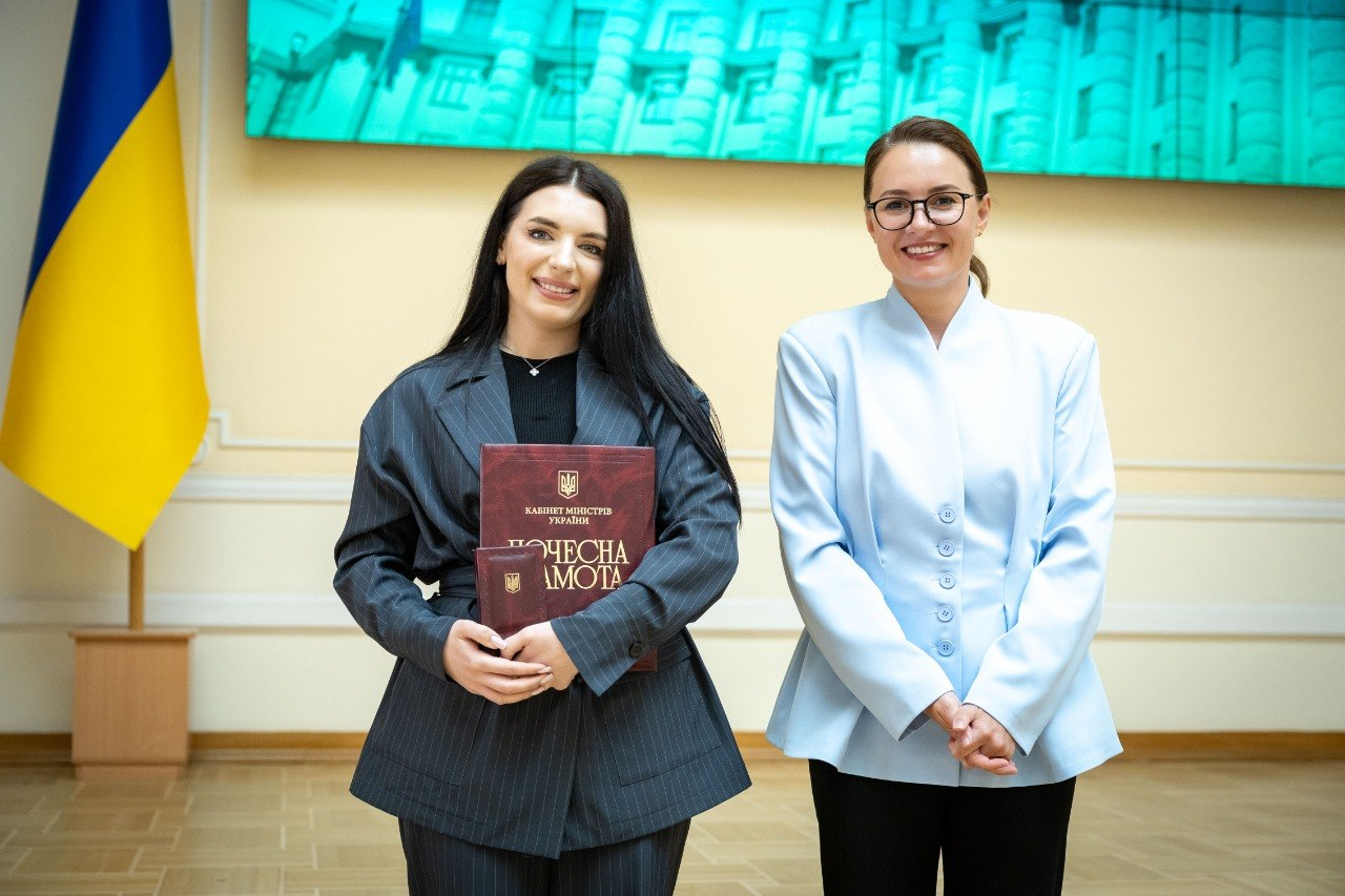 Жінка в темному костюмі в смужку з папкою «Почесна грамота» від Кабінету Міністрів України та жінка в окулярах і світло-блакитному жакеті посміхаються на тлі державного прапора України.