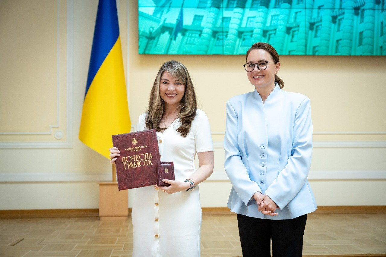 Жінка в білій сукні з папкою «Почесна грамота» від Кабінету Міністрів України та нагрудним знаком і жінка в окулярах та світло-блакитному жакеті посміхаються на тлі державного прапора України.