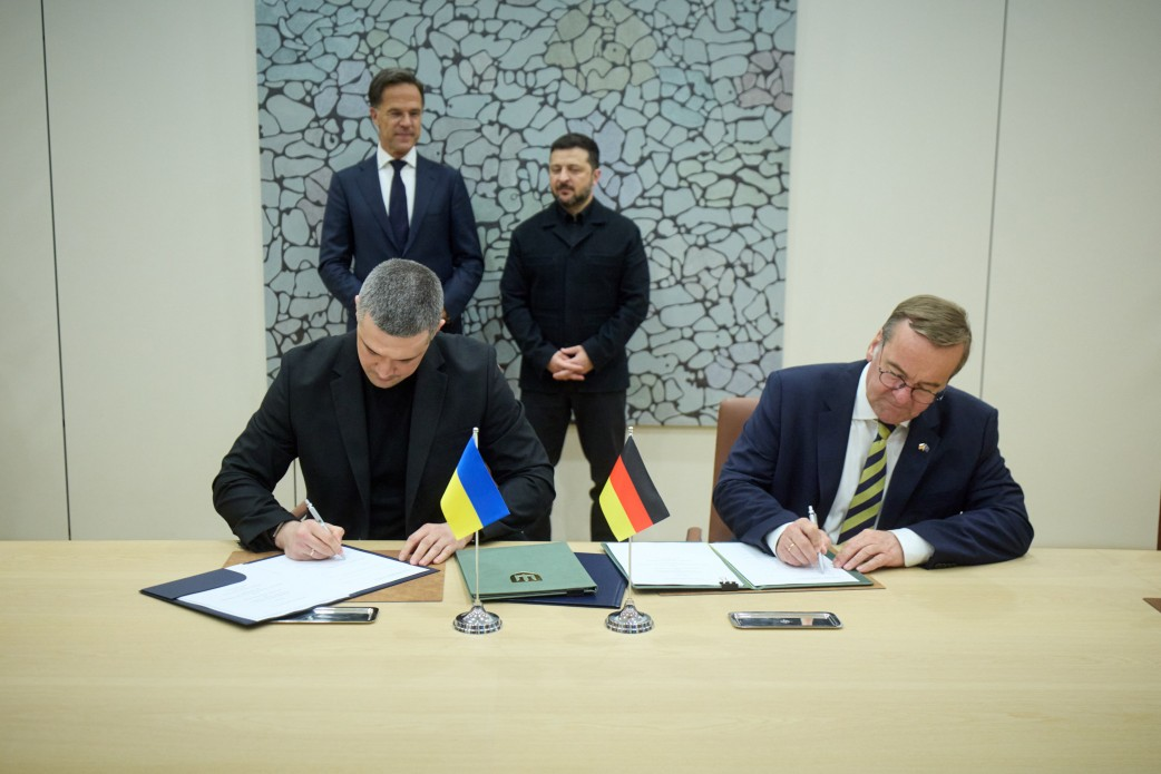 Два чоловіки у костюмах сидять за столом і підписують документи. Перед ними стоять маленькі настільні прапорці України та Німеччини. Позаду них стоять ще двоє чоловіків у діловому одязі та спостерігають за процесом підписання.