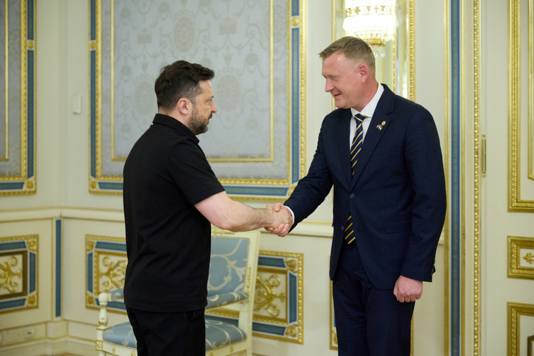 Двоє чоловіків тиснуть один одному руки під час офіційної зустрічі в класично оформленому кабінеті. Один чоловік одягнений у чорну сорочку поло, інший — у темно-синій діловий костюм із краваткою.