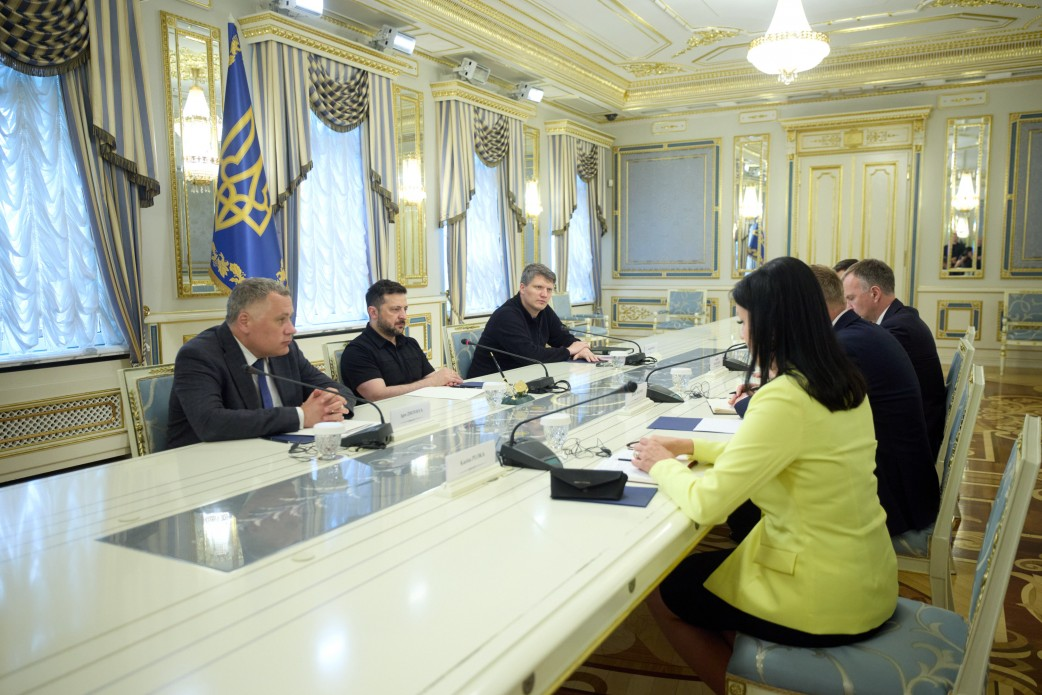Група людей у діловому та повсякденному одязі сидить за довгим білим столом під час переговорів. Зустріч проходить в офіційному залі із золотистим оздобленням, великими вікнами з гардинами та прапором України на задньому плані.