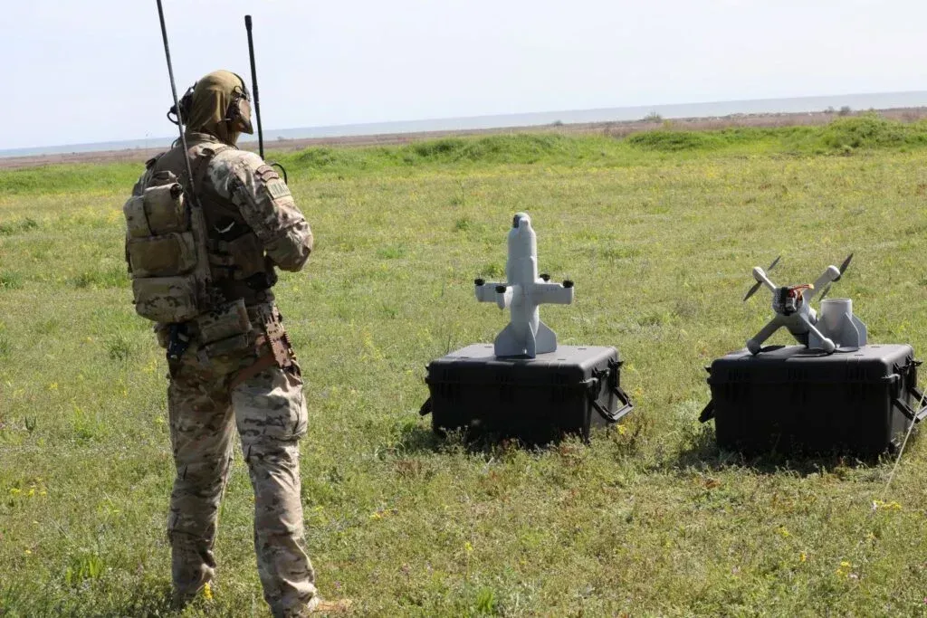 Військовий у камуфляжі та спорядженні стоїть спиною до камери на зеленому полі та керує безпілотниками. Праворуч від нього на двох чорних захисних кейсах розміщено два різні типи малих дронів вертикального зльоту.