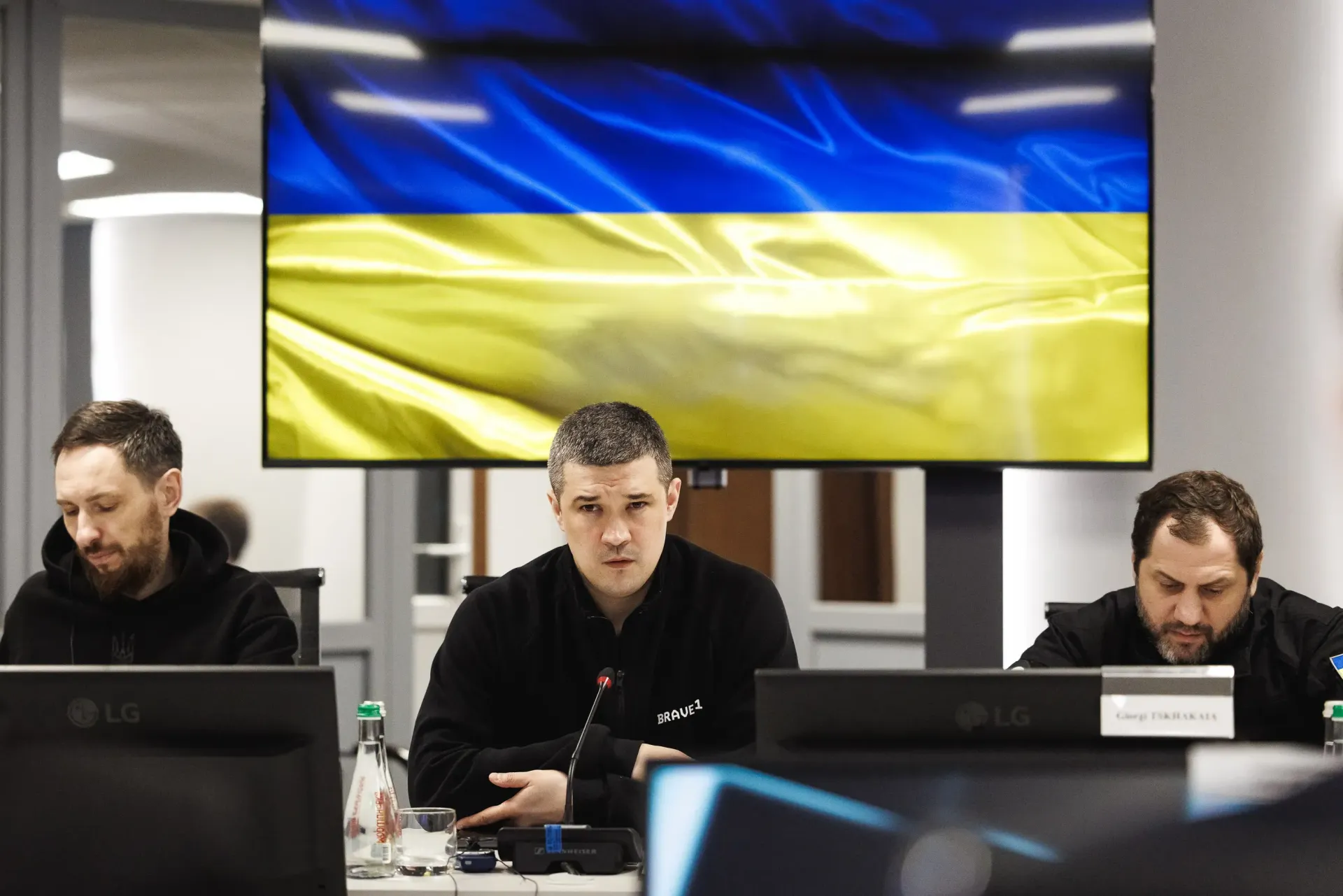 Троє чоловіків у темному одязі сидять за столом під час засідання. На великому екрані позаду них розгорнуто зображення державного прапора України.