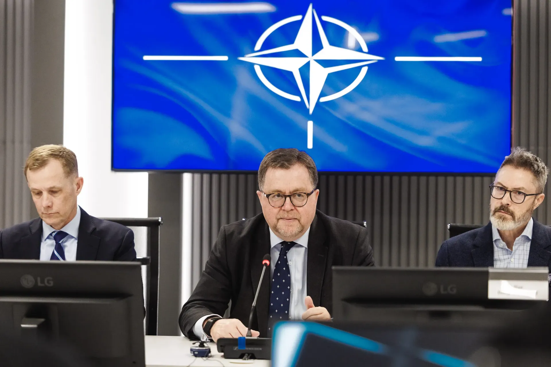 Троє чоловіків у ділових костюмах сидять за столом президії під час офіційної зустрічі. На великому синьому екрані позаду них зображено емблему НАТО.