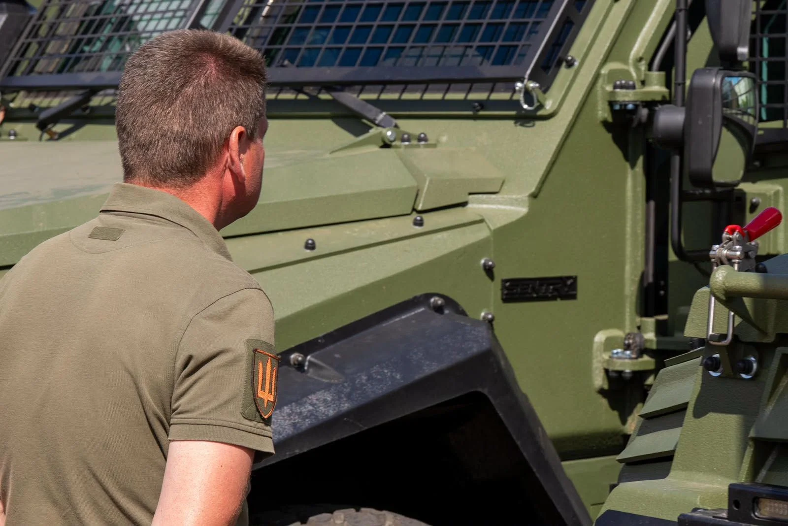 Чоловік у зеленій сорочці поло з нашивкою у формі українського тризуба на рукаві стоїть спиною до камери та оглядає зелений військовий бронеавтомобіль із захисною решіткою на лобовому склі.