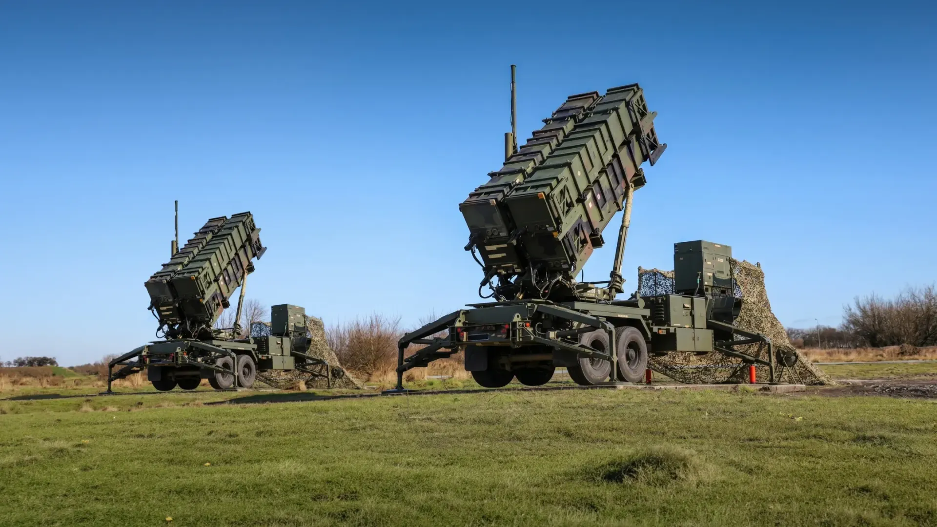 Дві пускові установки зенітно-ракетного комплексу Patriot розгорнуті на зеленому полі під безхмарним синім небом. Машини з піднятими під нахилом контейнерами для ракет розташовані поруч одна з одною на відкритій місцевості.