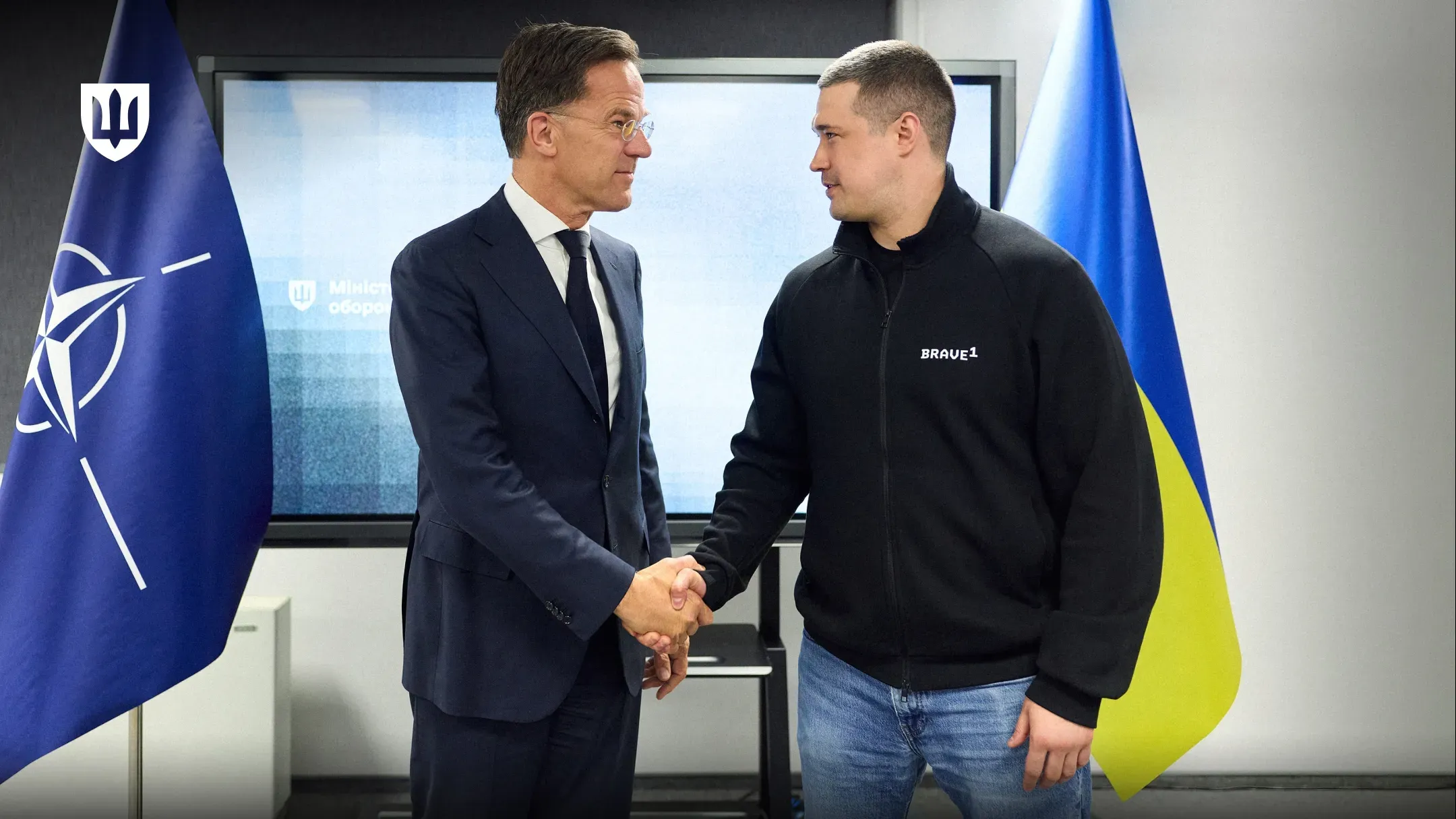 Двоє чоловіків тиснуть один одному руки, стоячи в кабінеті на тлі прапорів НАТО та України. Один чоловік одягнений у класичний діловий костюм із краваткою, а інший — у чорну спортивну кофту з написом «Brave1» та джинси.