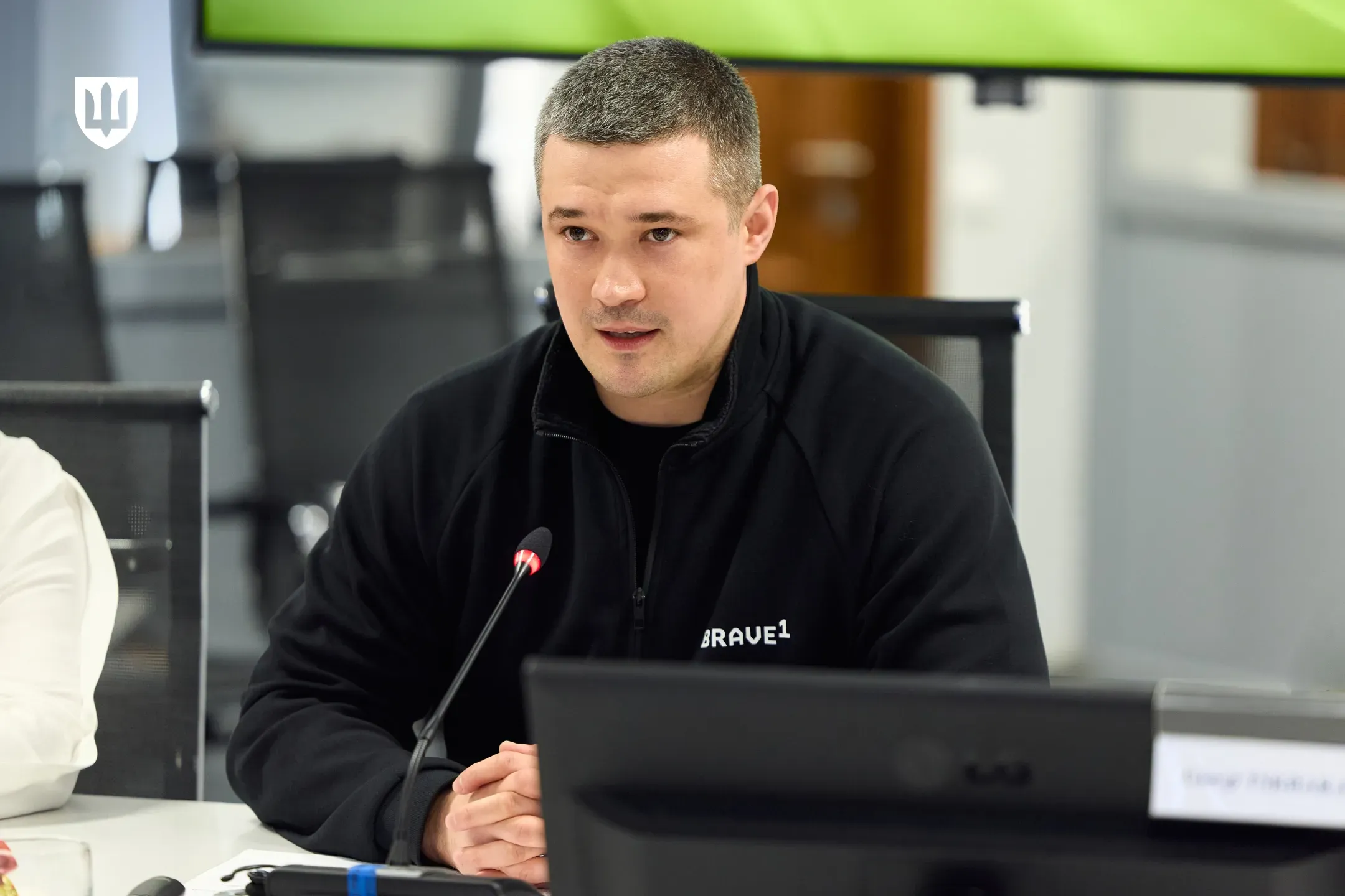 Чоловік із короткою стрижкою у чорній кофті з білим написом «Brave1» сидить за столом президії перед мікрофоном та монітором під час виступу чи робочої зустрічі. У лівому верхньому кутку зображення розміщено білий герб України у формі тризуба на щиті.