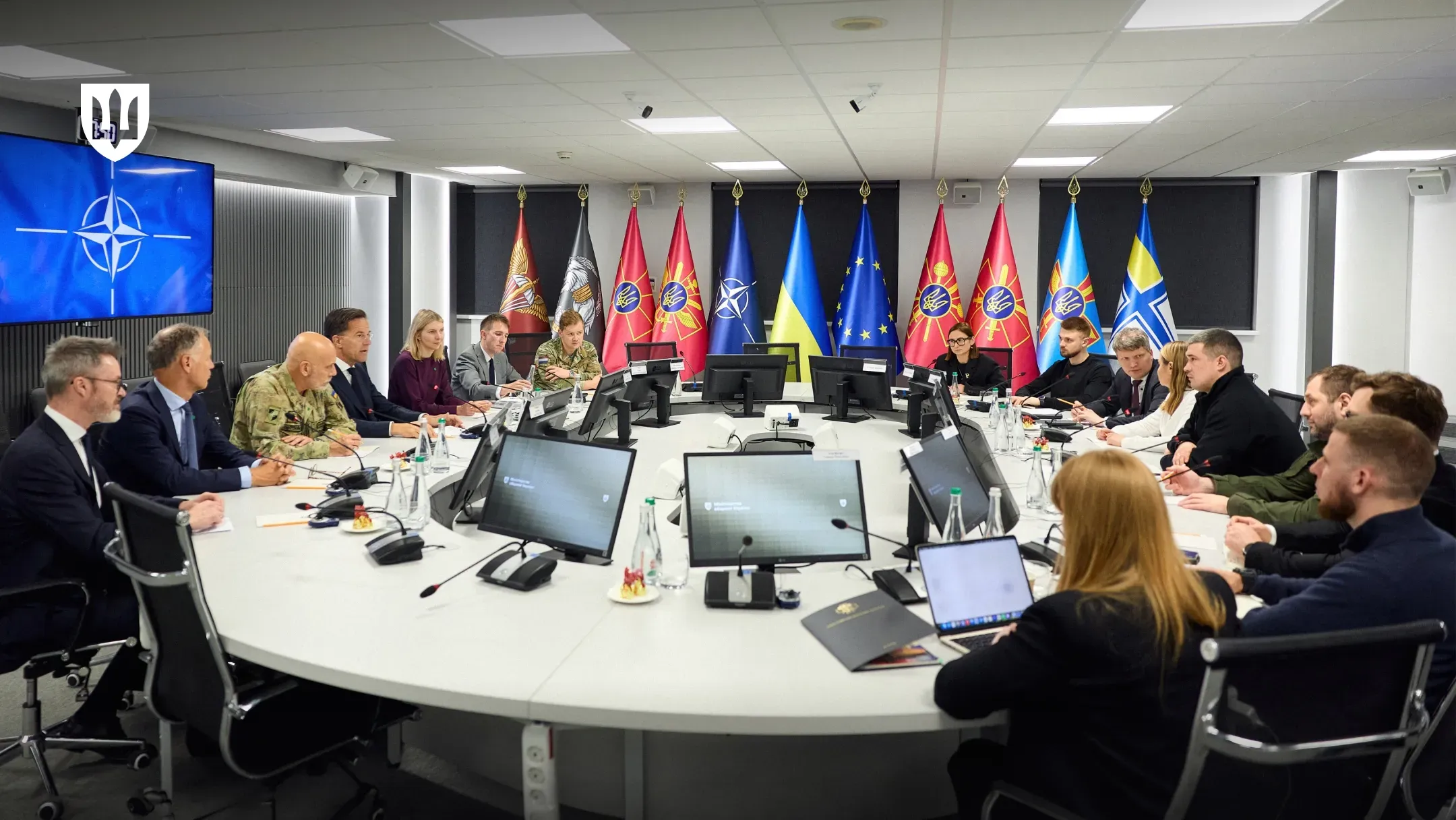 Велика група людей у цивільному одязі та військовій формі сидить навколо великого овального столу під час офіційних переговорів у конференц-залі. На столі встановлені комп'ютерні монітори та мікрофони, а на задньому плані розгорнуто ряд прапорів, серед яких прапори України, ЄС, НАТО та військових підрозділів.
