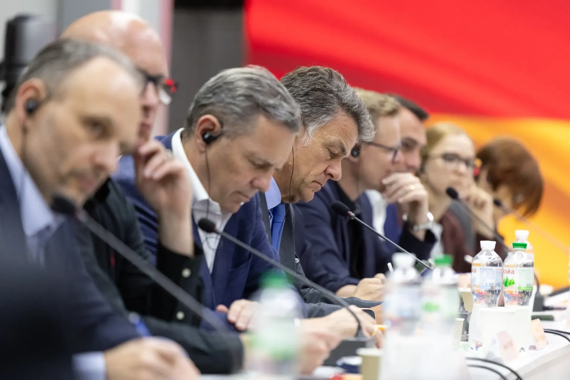 Люди у діловому одязі сидить за довгим столом президії під час офіційного заходу. Більшість із них слухають виступи у навушниках для синхронного перекладу, а на столі перед ними встановлено мікрофони та розставлено пляшки з водою.
