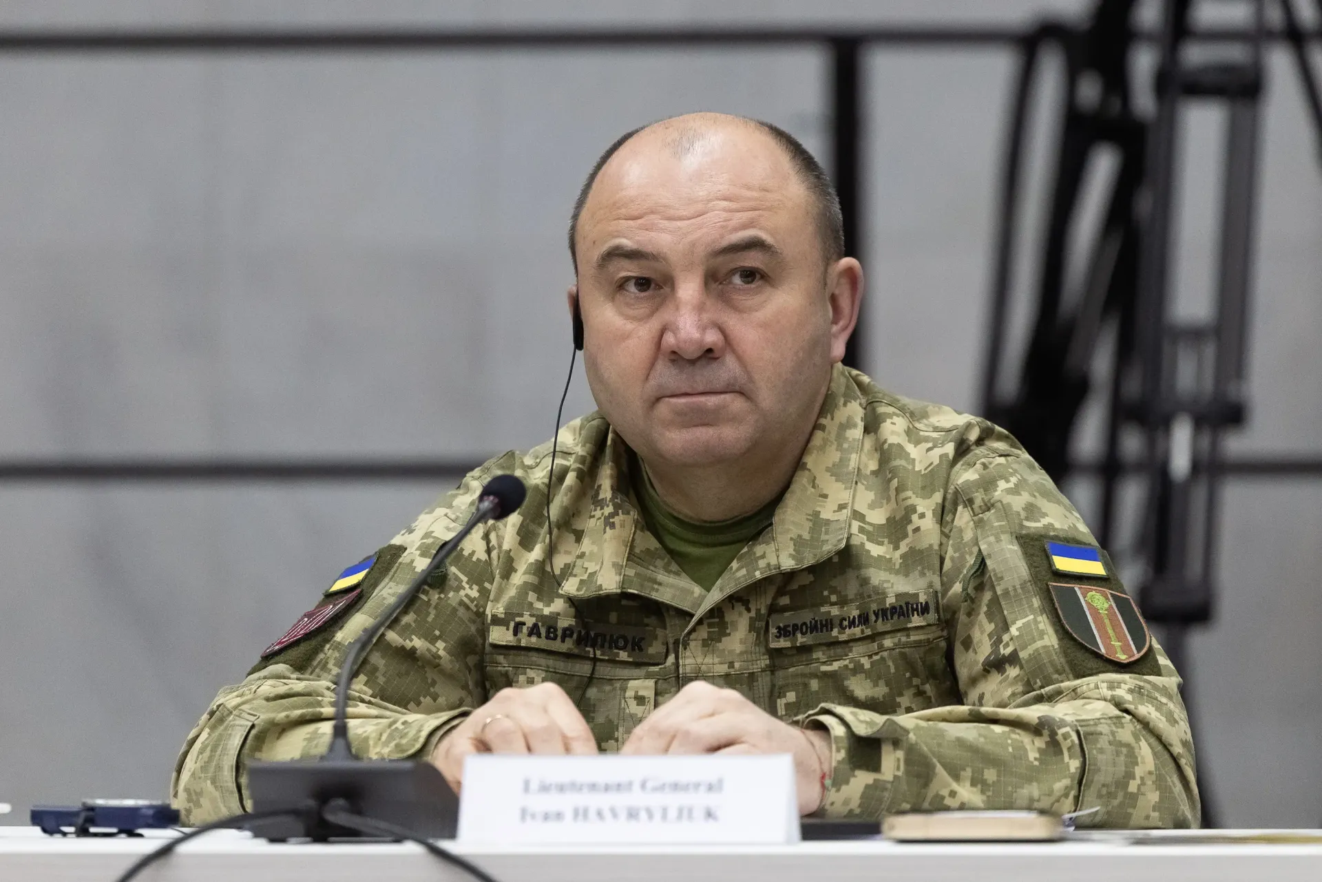 Чоловік у військовій камуфляжній формі із нашивками Збройних Сил України сидить за столом перед мікрофоном під час засідання.