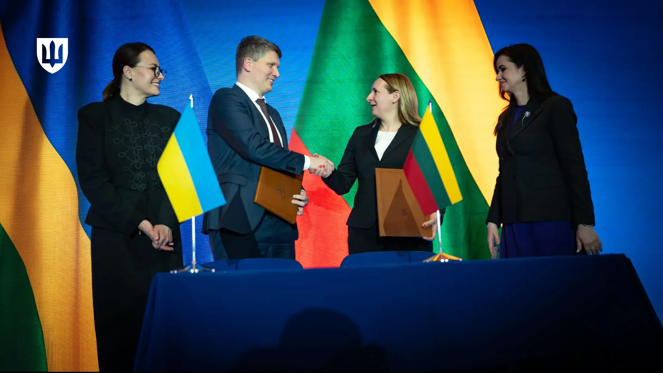 Чоловік та жінка тиснуть руки після підписання документів за столом, на якому стоять маленькі прапорці України та Литви. Обабіч них стоять ще дві жінки в діловому одязі, а на задньому плані розгорнуті великі державні прапори цих країн.